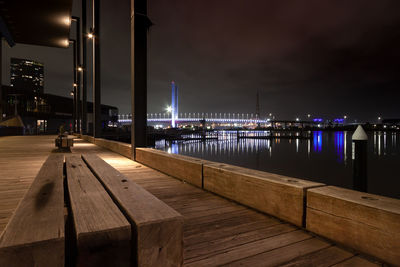 Illuminated pier and wooden bench over harbour against sky at night