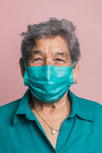 Smiling aged female using blue protective medical mask from coronavirus while looking at camera on pink background in studio