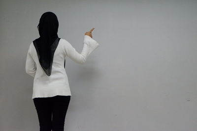 Rear view of woman in hijab pointing while standing against gray wall