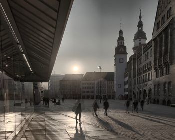 Blurred motion of people walking on city street amidst buildings against sky in city during sunset