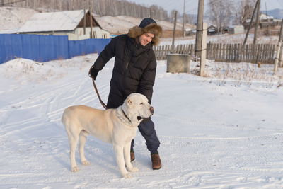 A young man stands with a big dog on a winter street in the background of a village house.