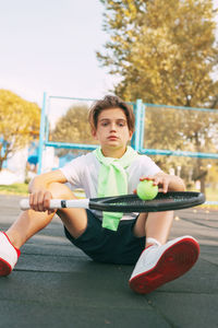 Portrait of boy with tennis racket sitting in court