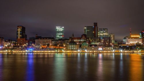 Illuminated buildings by river against sky at night