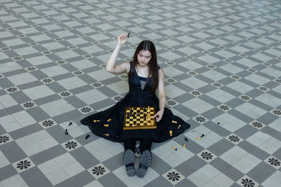 Beautiful girl in black dress plays chess in hall checkered floor