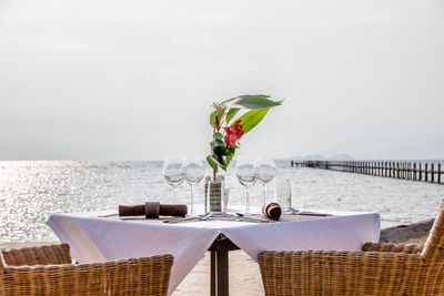 Flower vase and wineglasses on table by sea against sky