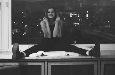 Portrait of happy woman sitting with legs apart on kitchen counter against window at home