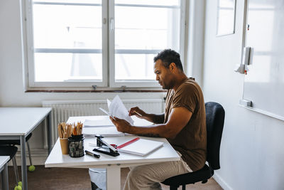 Focused male teacher checking test papers while sitting near table in classroom at junior high school