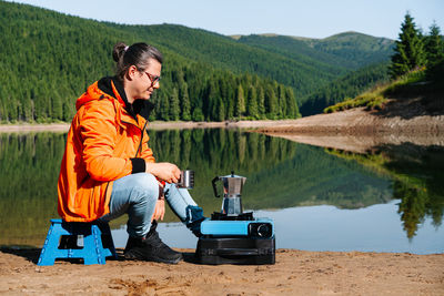 Side view of a man sitting by camping stove by the lake against mountains