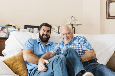 Portrait of adult grandson and his grandfather sitting on the couch at home tickling each other