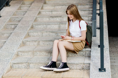 A cheerful teenage girl is sitting on the steps of the school, writing a message on her phone 