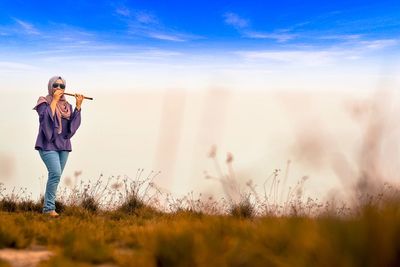 Young woman playing flute while standing on grassy field against cloudy sky