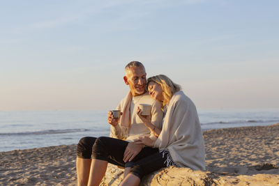 Couple having tea sitting on tree log at beach on sunset