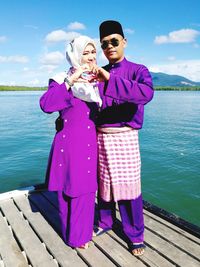 Full length of couple in purple traditional clothing making heart shape with hands by lake on jetty