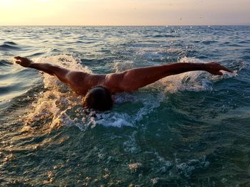 Shirtless young man with arms outstretched swimming in sea against sky during sunset