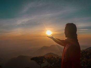 Optical illusion of man holding sun while standing against mountains and sky during sunset