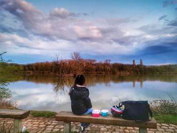 Rear view of woman sitting by lake against sky