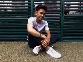 Smiling young man sitting on footpath against fence