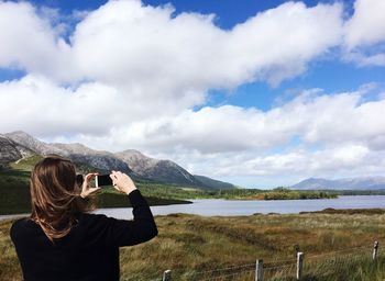 Rear view of woman photographing lake against sky