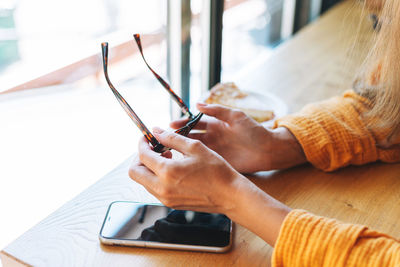 Crop photo of young woman holds sunglasses in hands with mobile phone on table in cafe