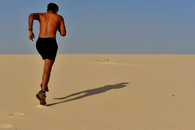 Full length of shirtless young man running on sand at desert during sunny day