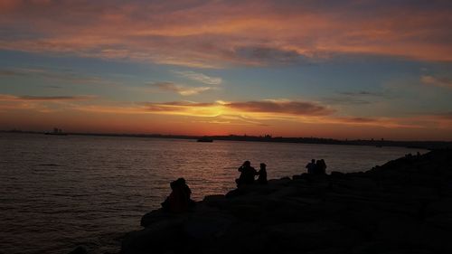 Silhouette people sitting by sea against sky during sunset