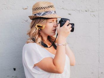 Side view of young woman wearing hat holding camera standing by wall outdoors