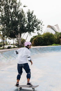 Full body of young ethnic person in casual outfit wearing protective helmet with knee pads and elbow pads riding skateboard in skate park
