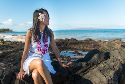 Young woman sitting on rock at beach against sky