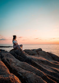 Man sitting on rock at beach against sky during sunset