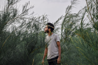 Young man looking away while standing on land against trees