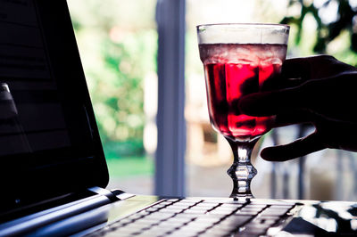 Cropped hand holding glass with red drink by laptop on table 