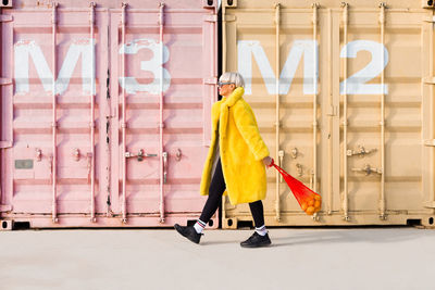 Side view of trendy female hipster in vibrant outfit walking along street with sack full of oranges and looking away