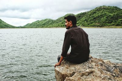 Young man sitting on rock in front of lake against cloudy sky