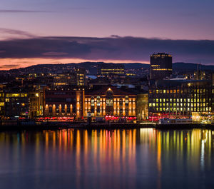 Illuminated buildings by river against sky at sunset