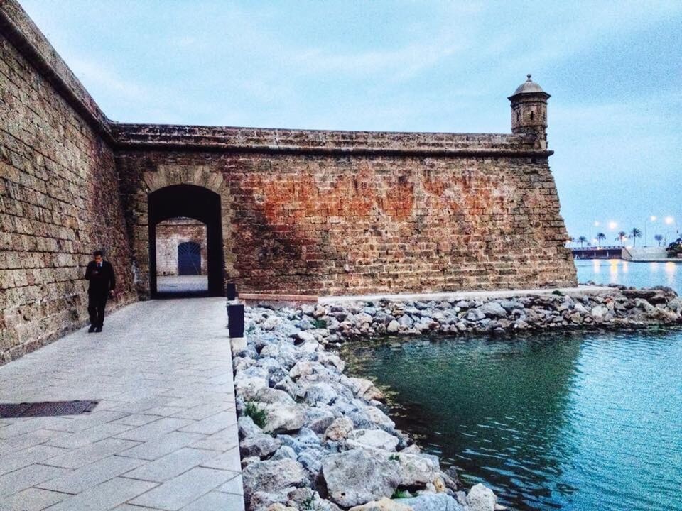 architecture, built structure, building exterior, water, sky, lighthouse, sea, day, cloud, lifestyles, men, blue, cloud - sky, walking, outdoors, incidental people, history, stone wall