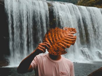 Young man covering face with leaf against waterfall in forest during autumn