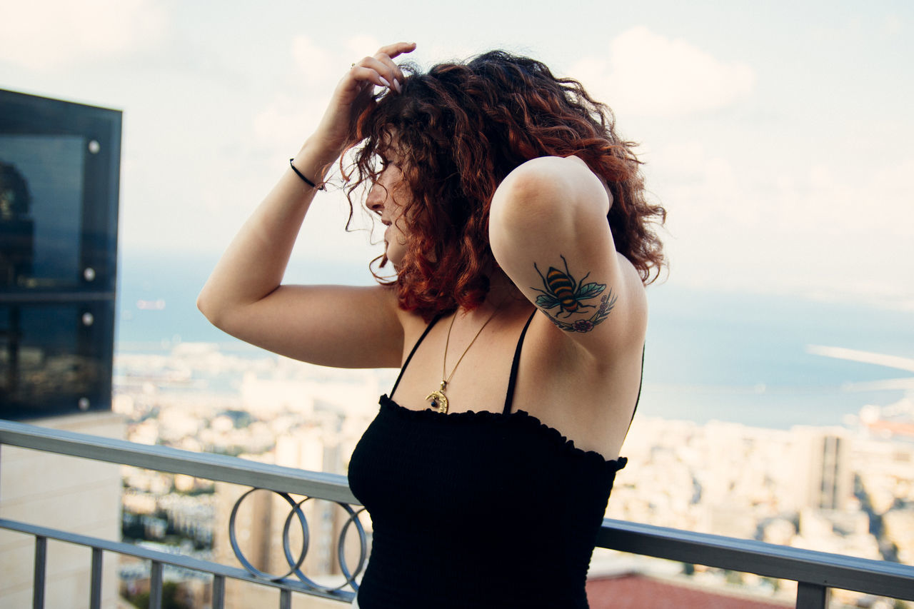 one person, real people, young women, young adult, leisure activity, focus on foreground, hairstyle, railing, lifestyles, standing, architecture, portrait, sky, hair, women, built structure, day, adult, brown hair, beautiful woman, balcony, outdoors, wind