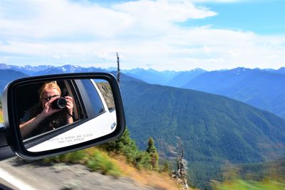 Reflection of woman photographing on side-view mirror against mountains