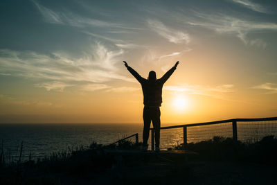 Silhouette man with arms raised standing in front of sea at sunset