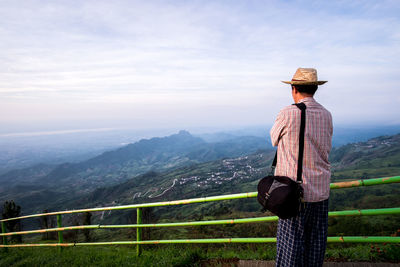 Rear view of man standing by railing on mountain against cloudy sky