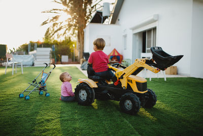 A baby girl and a toddler sitting on a toy tractor in the yard