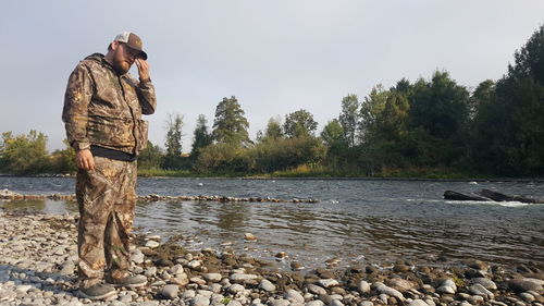 Side view of mid adult man in camouflage clothing standing on rock by 
lake against sky