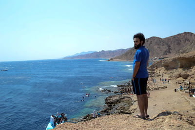  young man standing on cliff by beach against clear blue sky