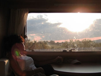 View of woman sitting by window at sunset