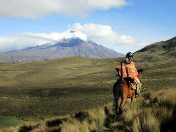 Person wearing a traditional poncho rides horseback across the paramo w/ volcano cotopaxi in view
