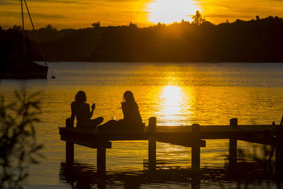 Silhouette people sitting on jetty against sea during sunset