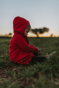Side view of girl sitting on grassy field during sunset