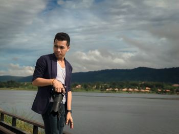 Man holding camera while looking away by lake against cloudy sky