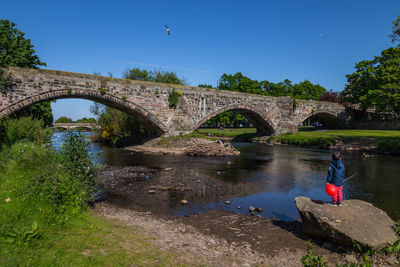 Boy standing on rock at lakeshore against arch bridge