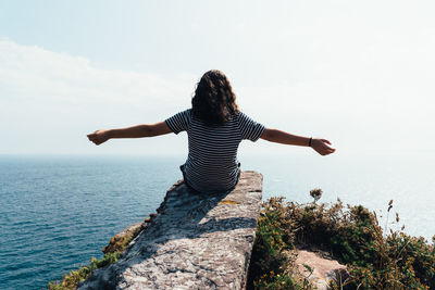 Rear view of girl with arms outstretched sitting on rock by sea against sky